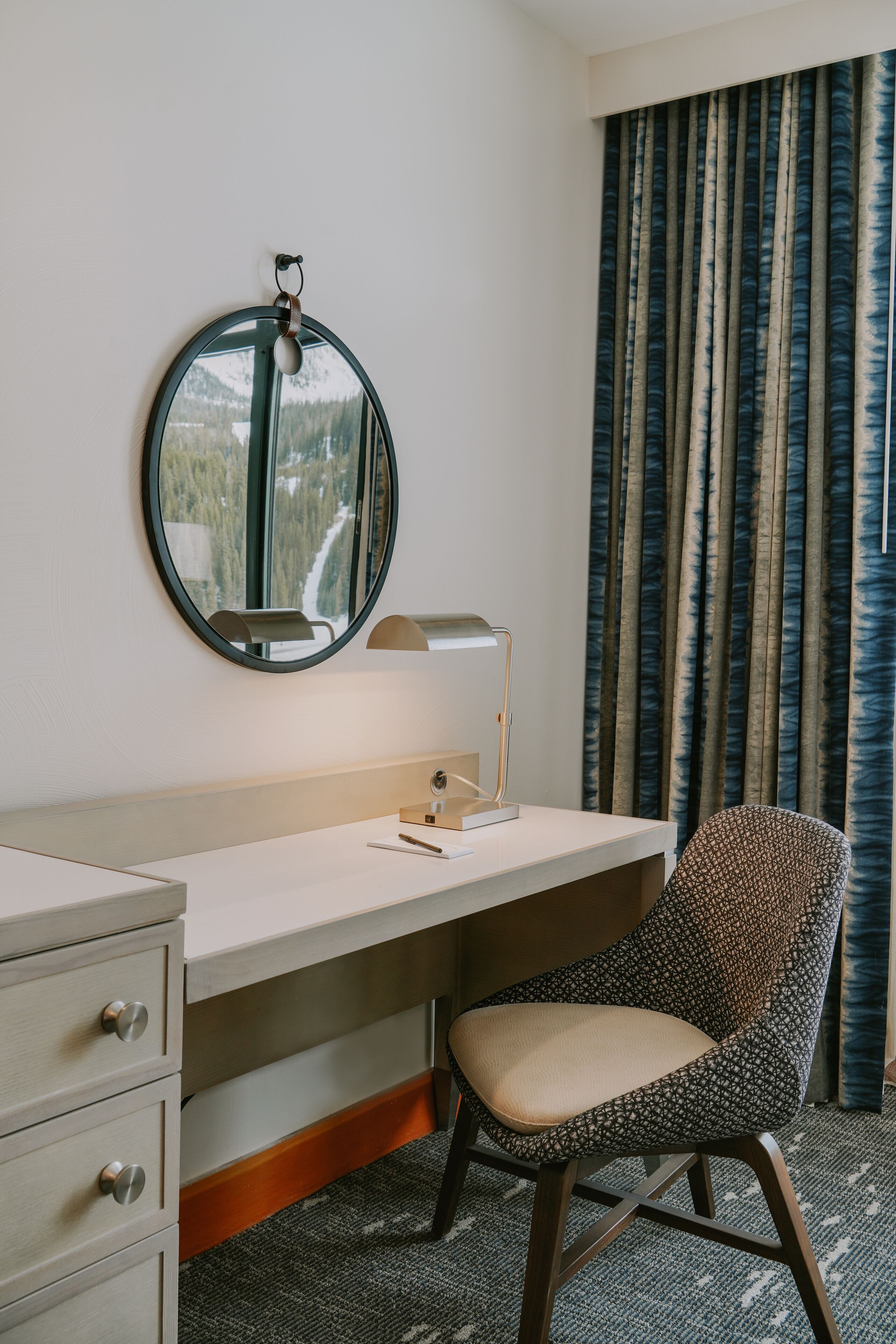 Habitación de hotel con un espejo redondo, un escritorio con lámpara, una silla y vistas a la montaña a través de la ventana.