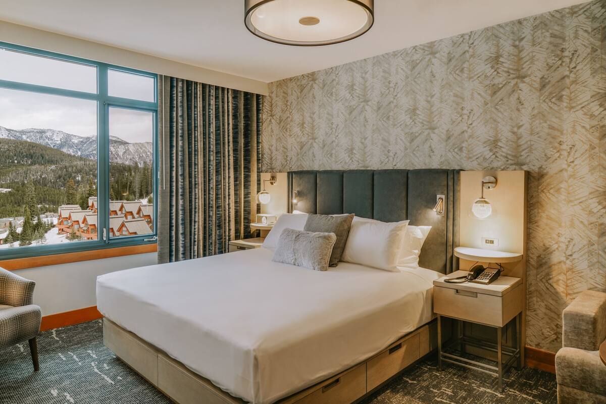 Ein Hotelzimmer mit einem großen Bett, zwei Nachttischen mit Lampen, einem Sessel und Blick auf Berge, sowie einer Wand mit Muster.