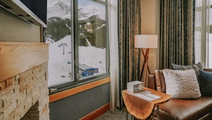 Une pièce dotée d’une grande fenêtre donnant sur un paysage montagneux enneigé, d’un mur en bois, d’un canapé en cuir marron agrémenté de coussins et d’une petite table en bois sur laquelle se trouve une lampe.