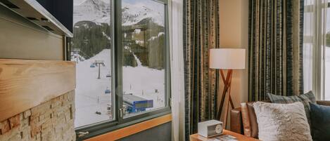 A room with a large window overlooking a snowy mountain landscape, a wooden wall, a brown leather sofa with cushions, and a small wooden table with a lamp.