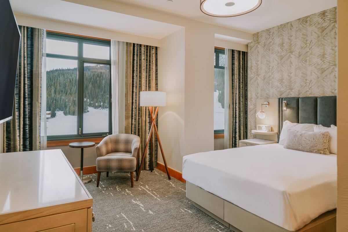 Ein Hotelzimmer mit einem großen Bett, einem Sessel, einem Schreibtisch, einer Lampe und Blick auf einen verschneiten Wald durch das Fenster.