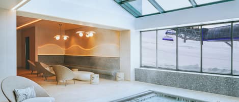 Een modern binnenzwembad met glazen dak, zitplaatsen en uitzicht op een skigebied.