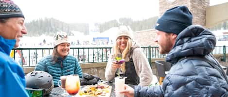 A group of people sitting at an outdoor table with food and drinks, with a snowy landscape and a clock tower in the background.