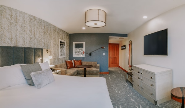 Habitación de hotel con cama, sofá, escritorio, cómoda, televisor y pasillo que conduce a otra habitación.