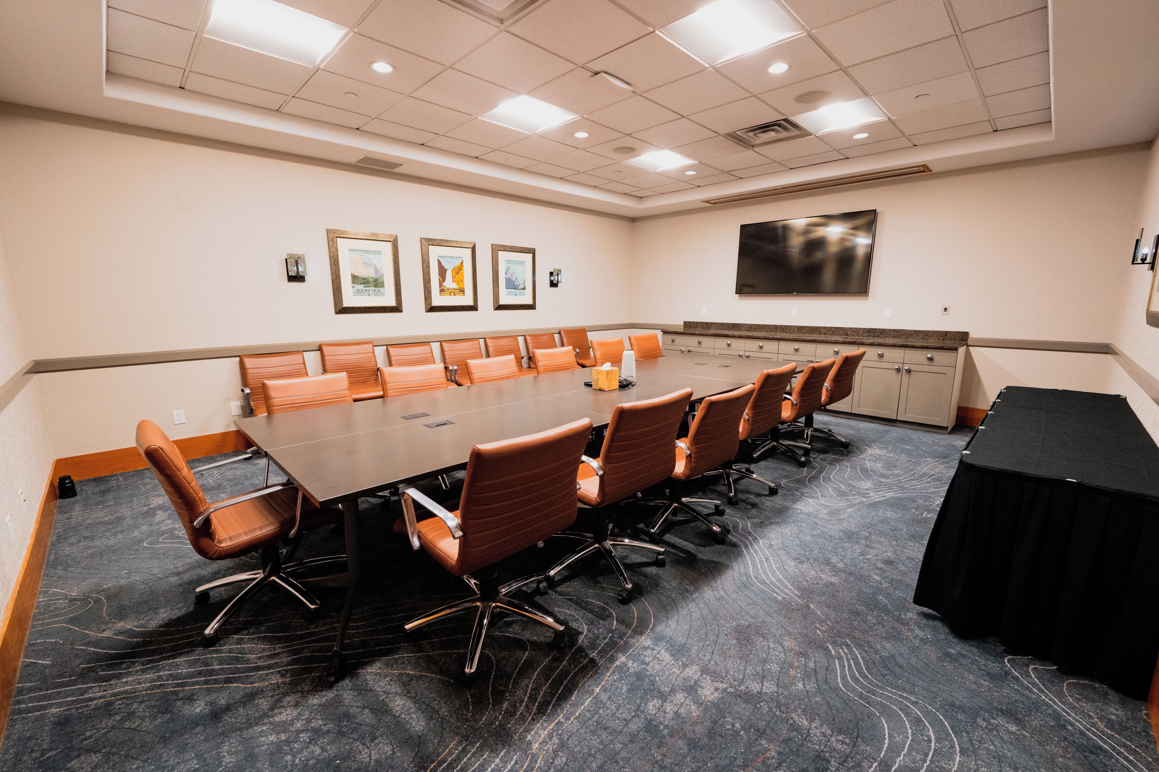 A conference room with a long table, multiple chairs, a flat-screen TV, framed pictures on the wall, and a carpeted floor.