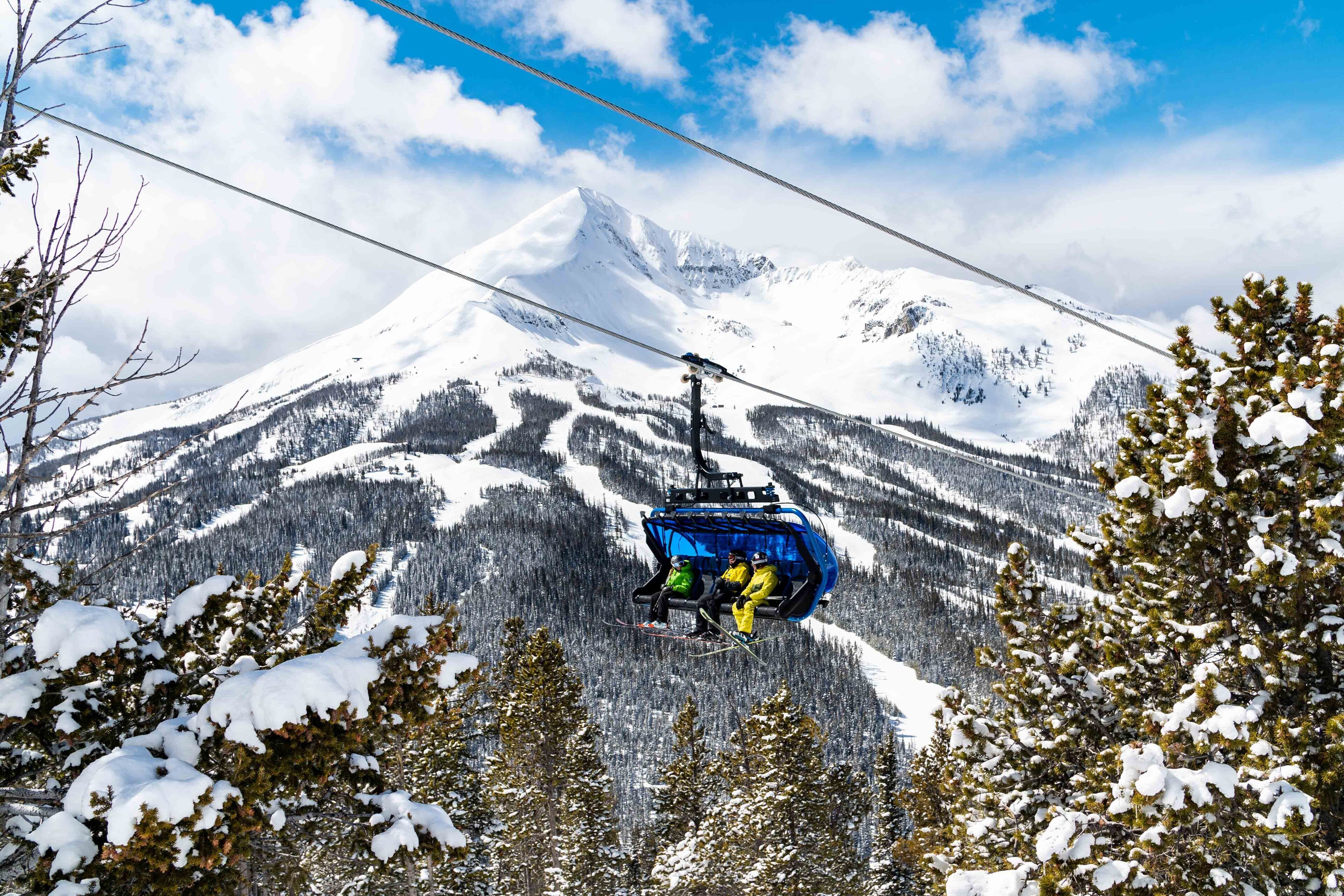 Eine Sesselbahn in einer verschneiten Berglandschaft mit Bäumen und blauem Himmel.