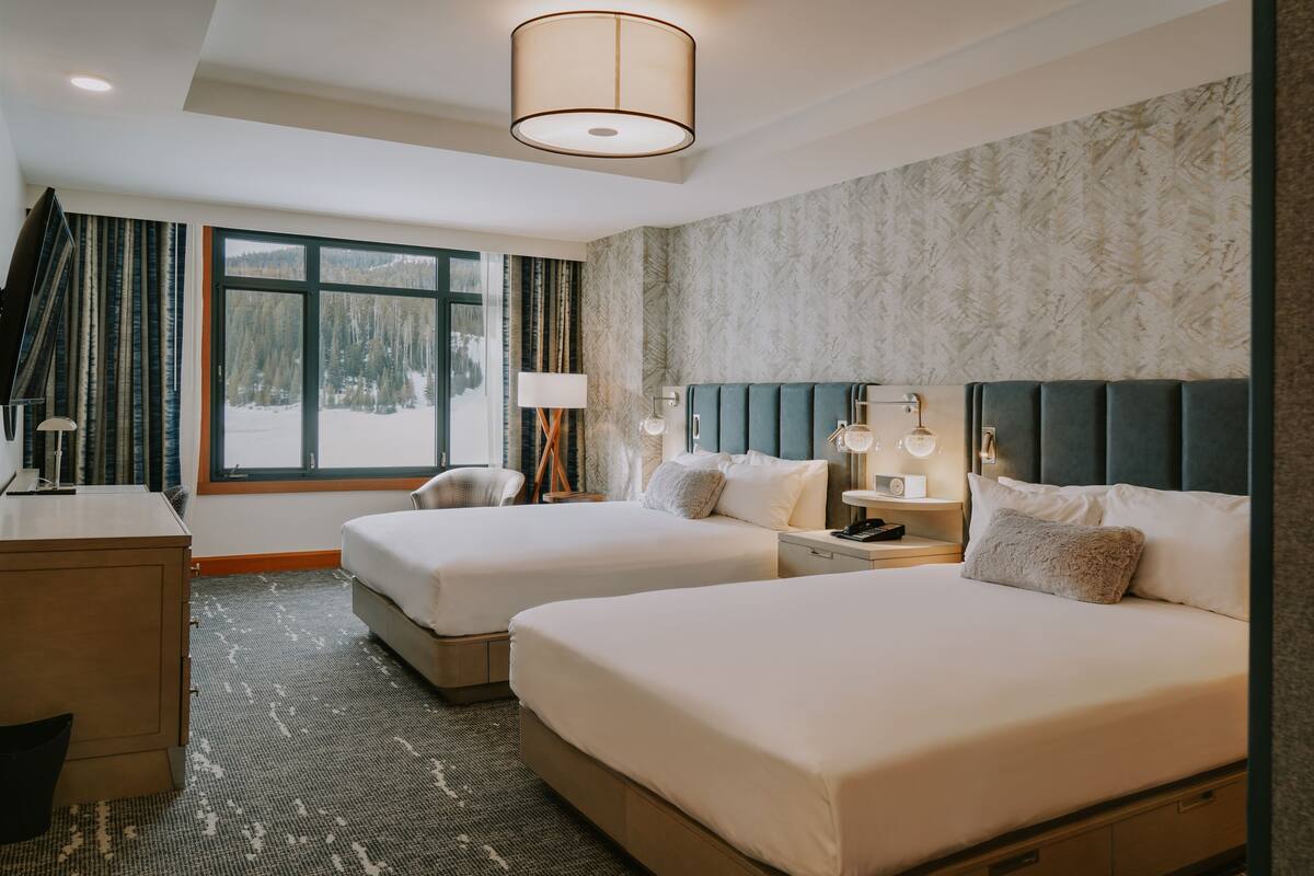 Ein Hotelzimmer mit zwei Betten, einem Schreibtisch, einem Stuhl, einer Lampe und Blick auf einen verschneiten Wald durch große Fenster.