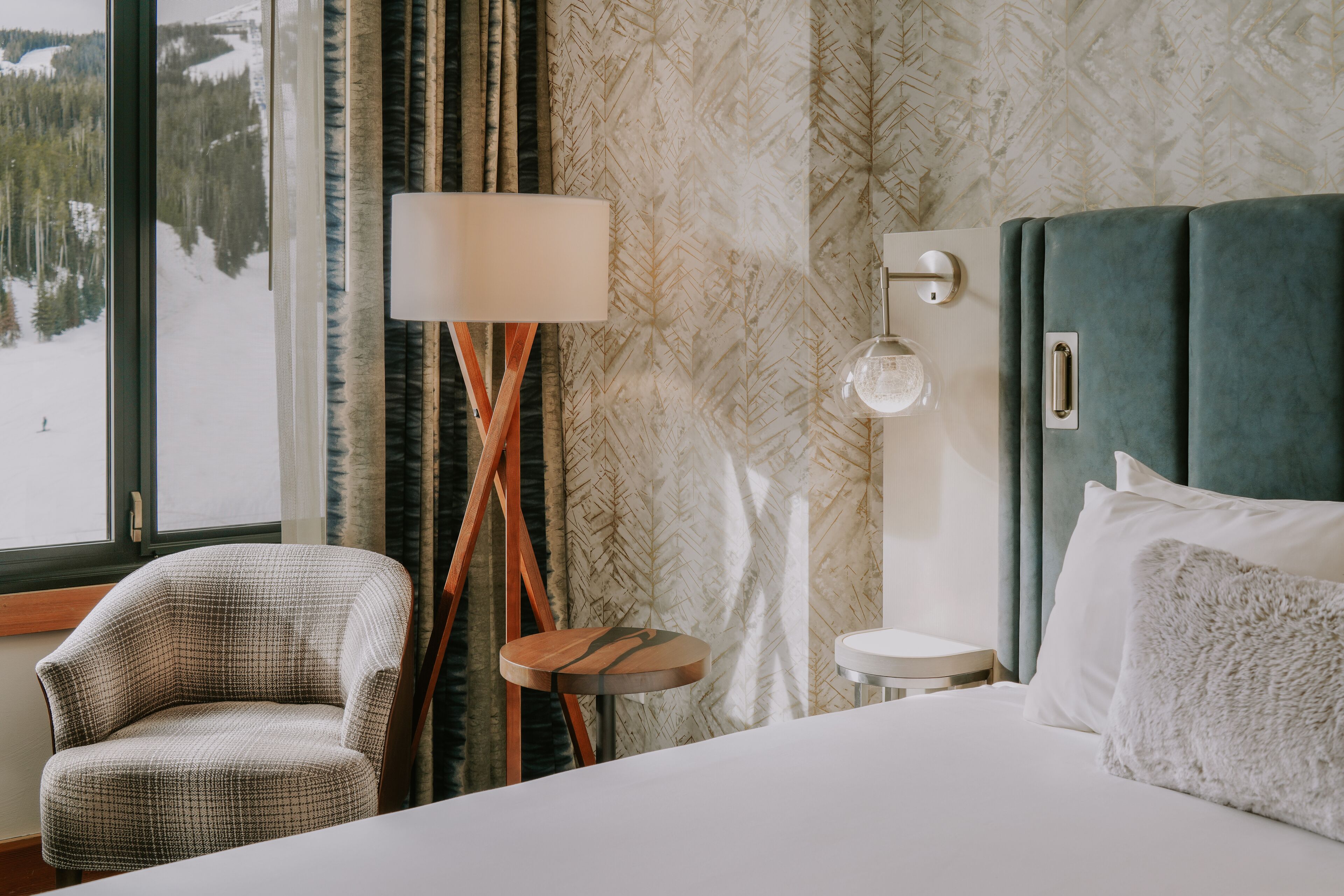 A hotel room with a bed, a chair, a small table, a lamp, and a view of a snowy landscape through a window.