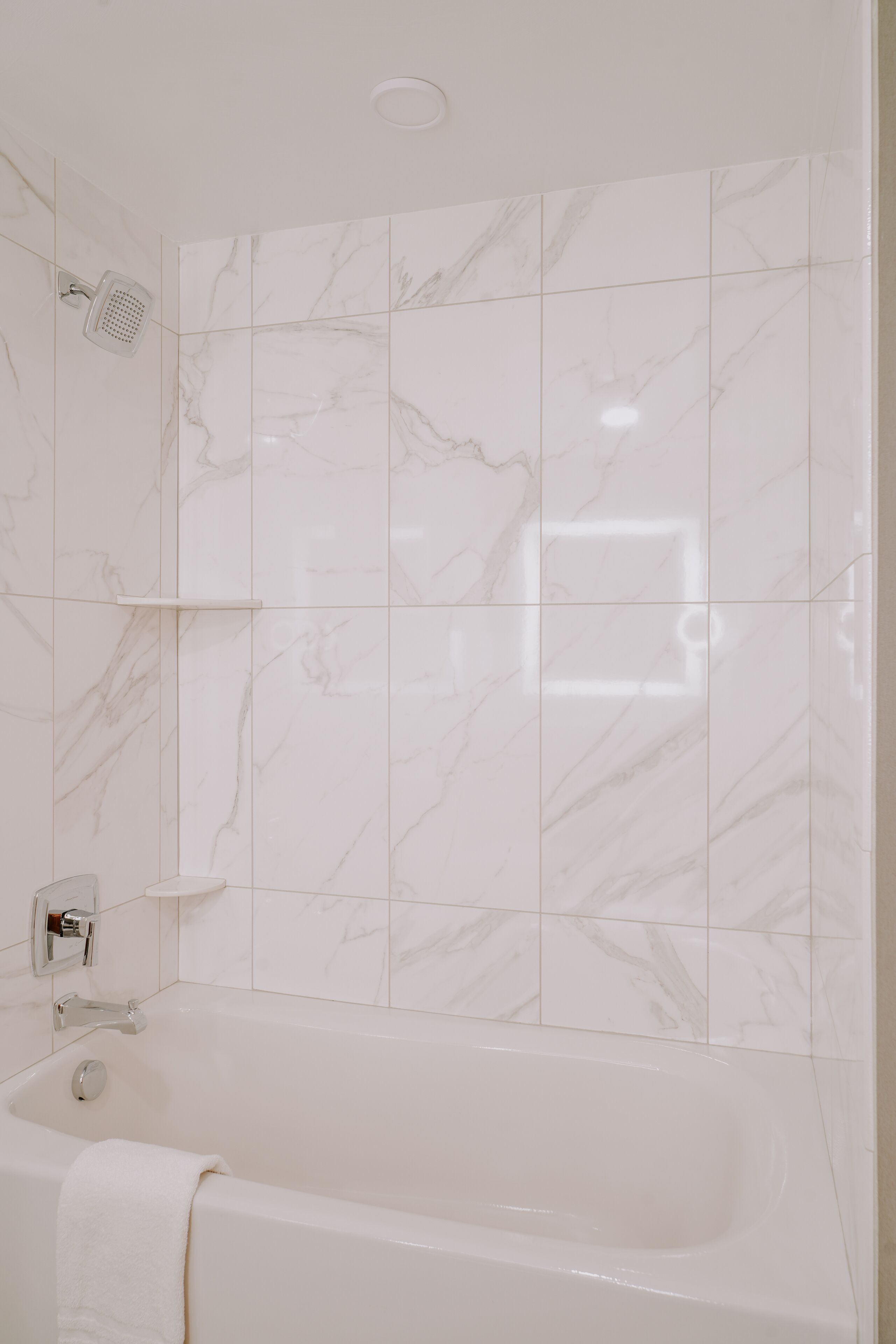 Une salle de bain avec un mur de douche recouvert de carreaux de marbre, une étagère intégrée et une baignoire avec une serviette.