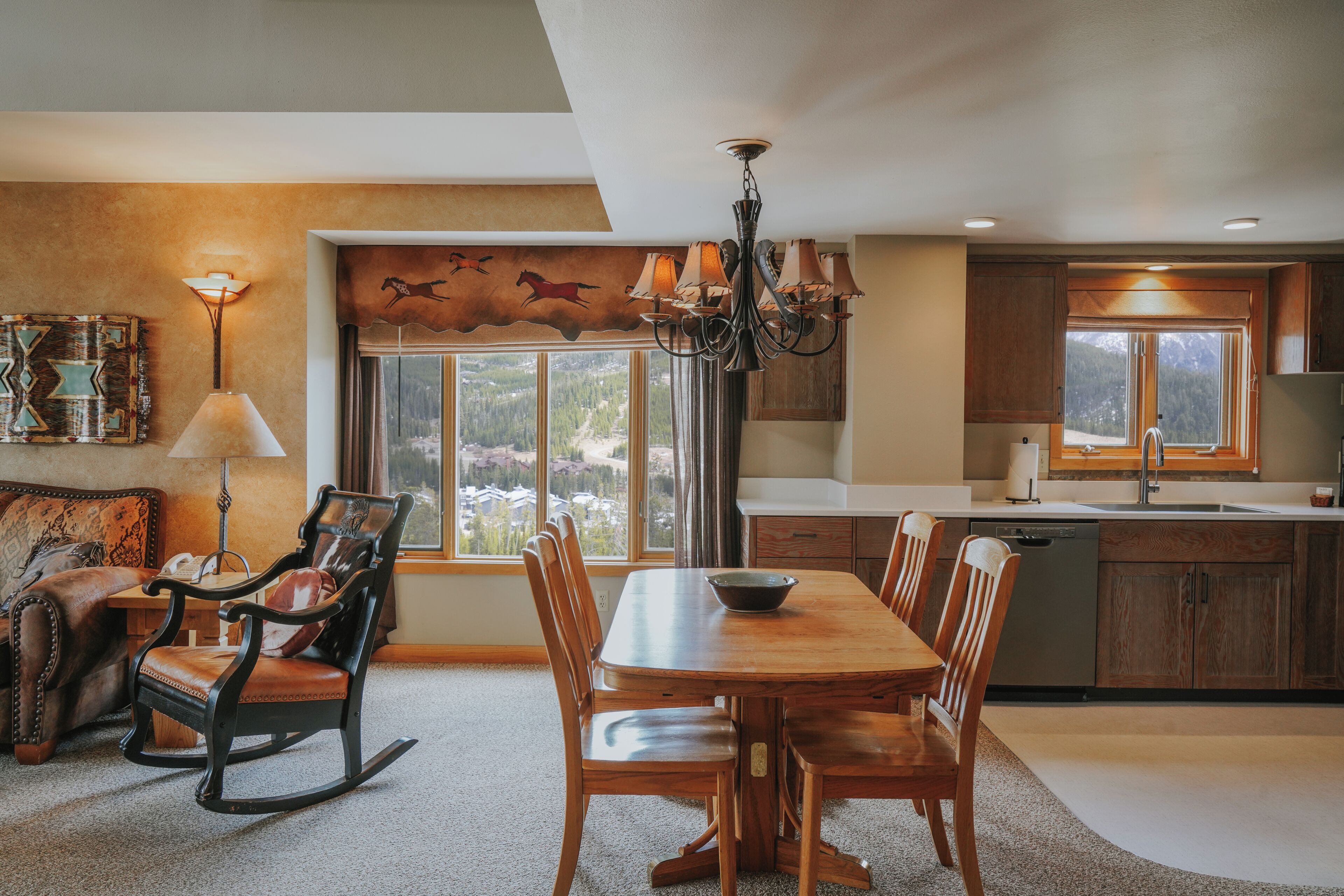 A dining area with a wooden table and chairs, a chandelier, and a view of the outdoors through large windows.