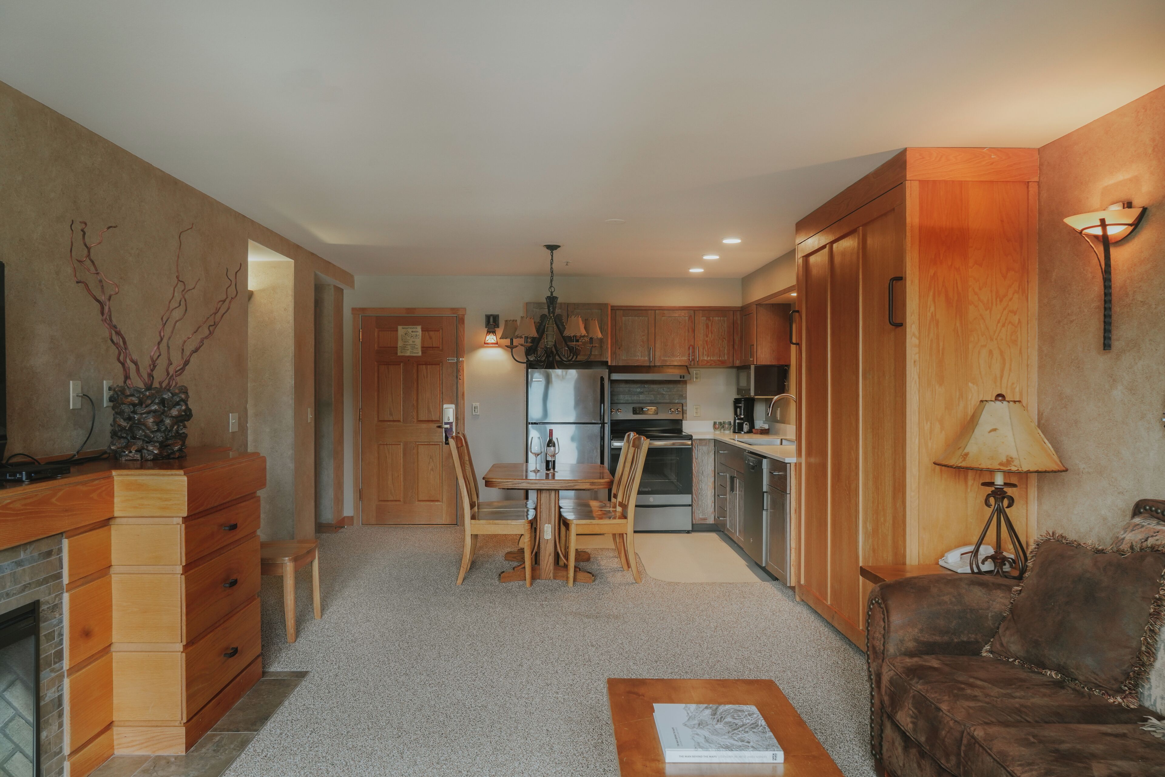 A modern kitchen with wooden cabinets, a dining table, and chairs. There is a fireplace, a coffee machine, and a lamp on the wall.