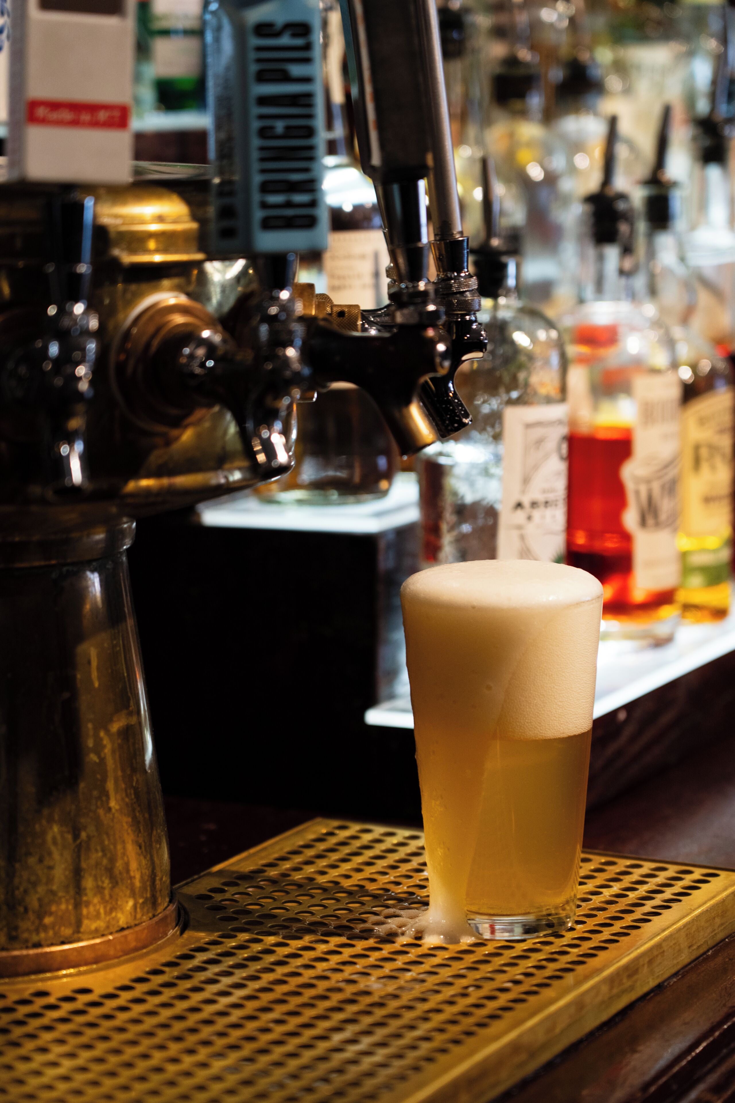 A bar with a tap dispensing a beverage into a glass, a metal coffee pot, and various bottles in the background.