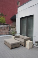 An outdoor seating area with a wicker sofa and ottoman, a potted plant, and a brick wall in the background.