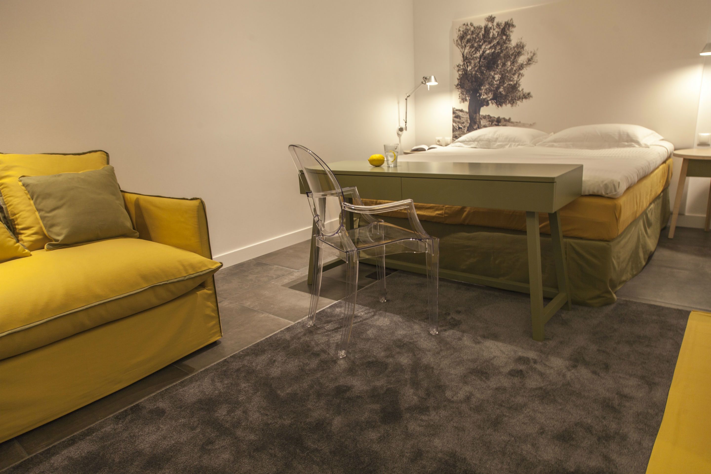 A room with a yellow sofa, a clear chair, a bed with white bedding, a desk, and a lamp.