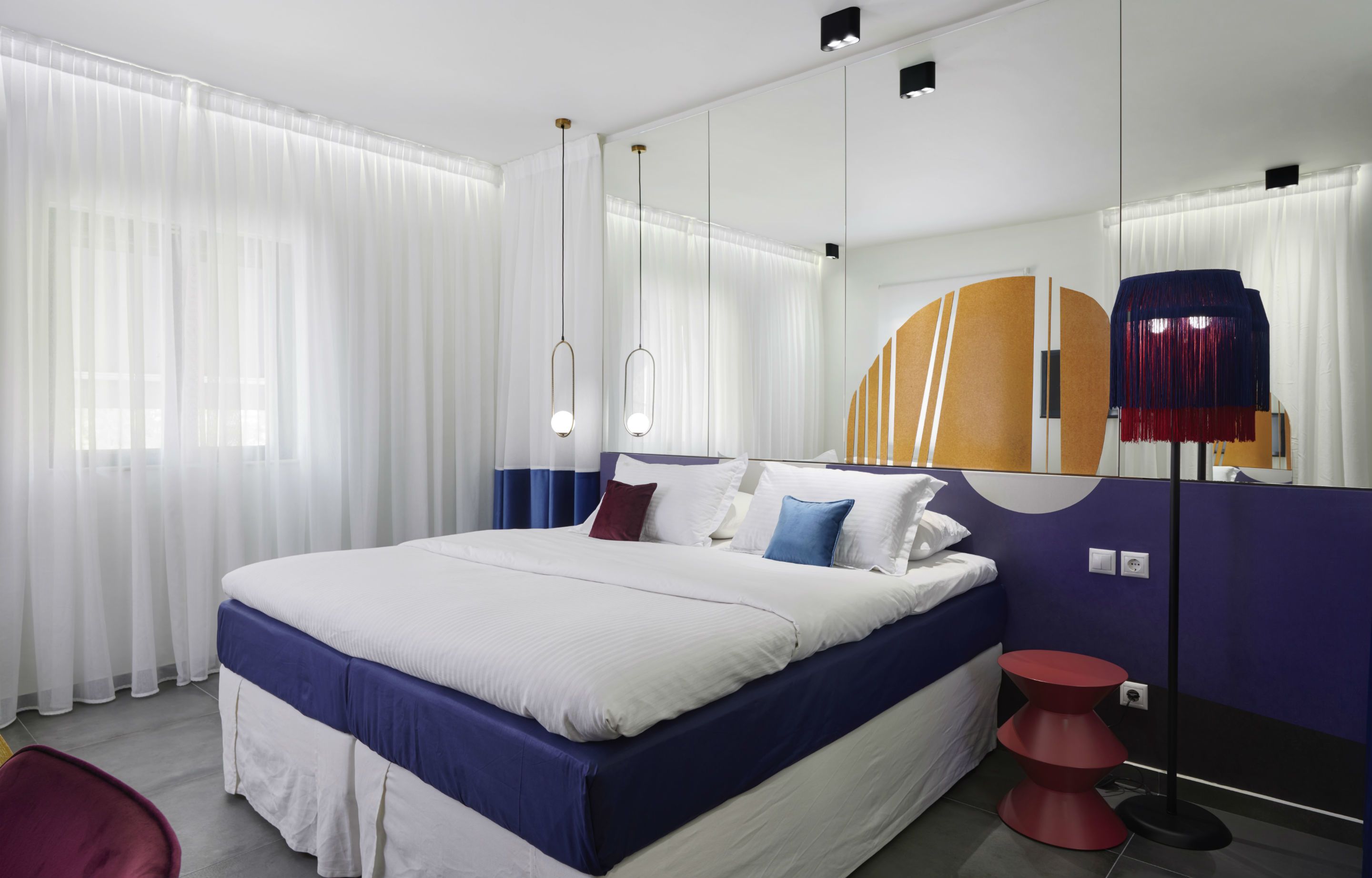 A modern hotel room with a large bed, white curtains, a red chair, and a unique wall design.