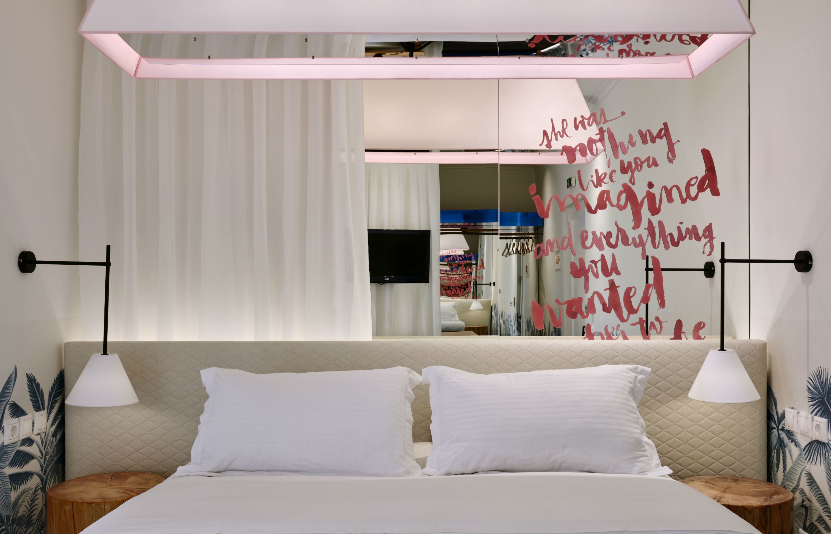 A modern bedroom with a bed, two bedside tables, a wall-mounted TV, and a decorative quote.