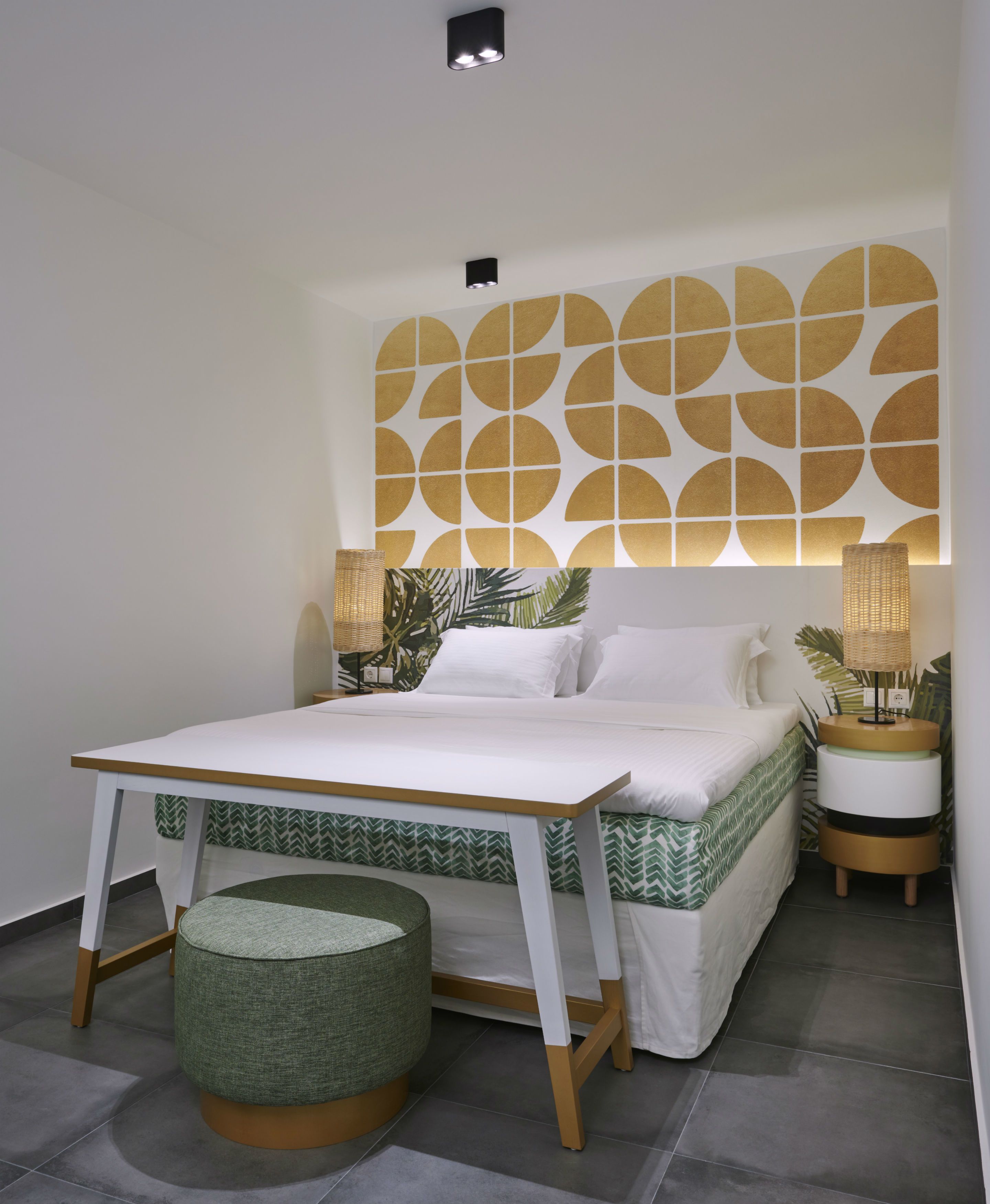 Un dormitorio moderno con una cama, mesitas de noche, lámparas de noche, un pequeño taburete redondo y un diseño geométrico en la pared.