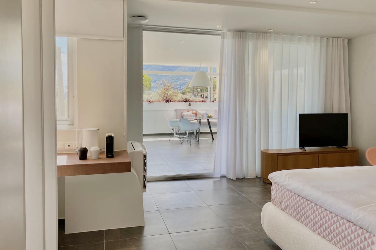 A modern hotel room with a bed, a TV, a desk with a lamp, and a view of the outdoors.