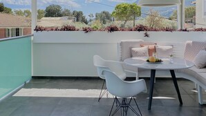 Un área de estar exterior moderna con una mesa y sillas blancas, una lámpara colgante grande en blanco y vistas a un paisaje montañoso a través de amplios ventanales.