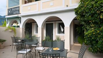 An outdoor seating area with tables and chairs, a sign for 'thomas BOUTIQUE HOTEL', and a green hedge.