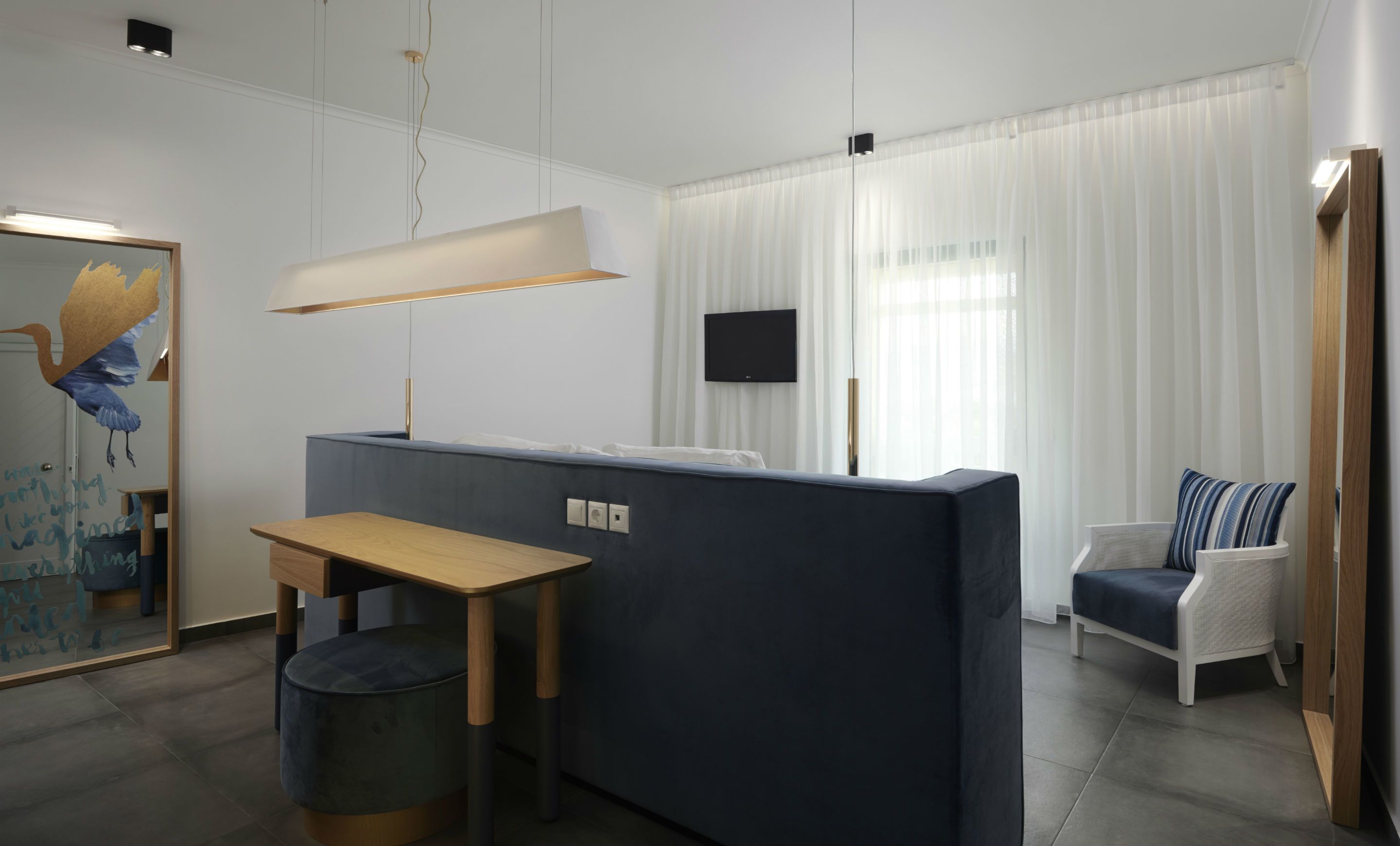 Een moderne woonkamer met een donkerblauwe bank, een houten tafel en een witte fauteuil. Een flatscreen televisie is aan de muur bevestigd en er is een groot raam met dunne gordijnen.