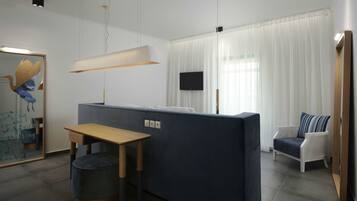 Een moderne woonkamer met een donkerblauwe bank, een houten tafel en een witte fauteuil. Een flatscreen televisie is aan de muur bevestigd en er is een groot raam met dunne gordijnen.