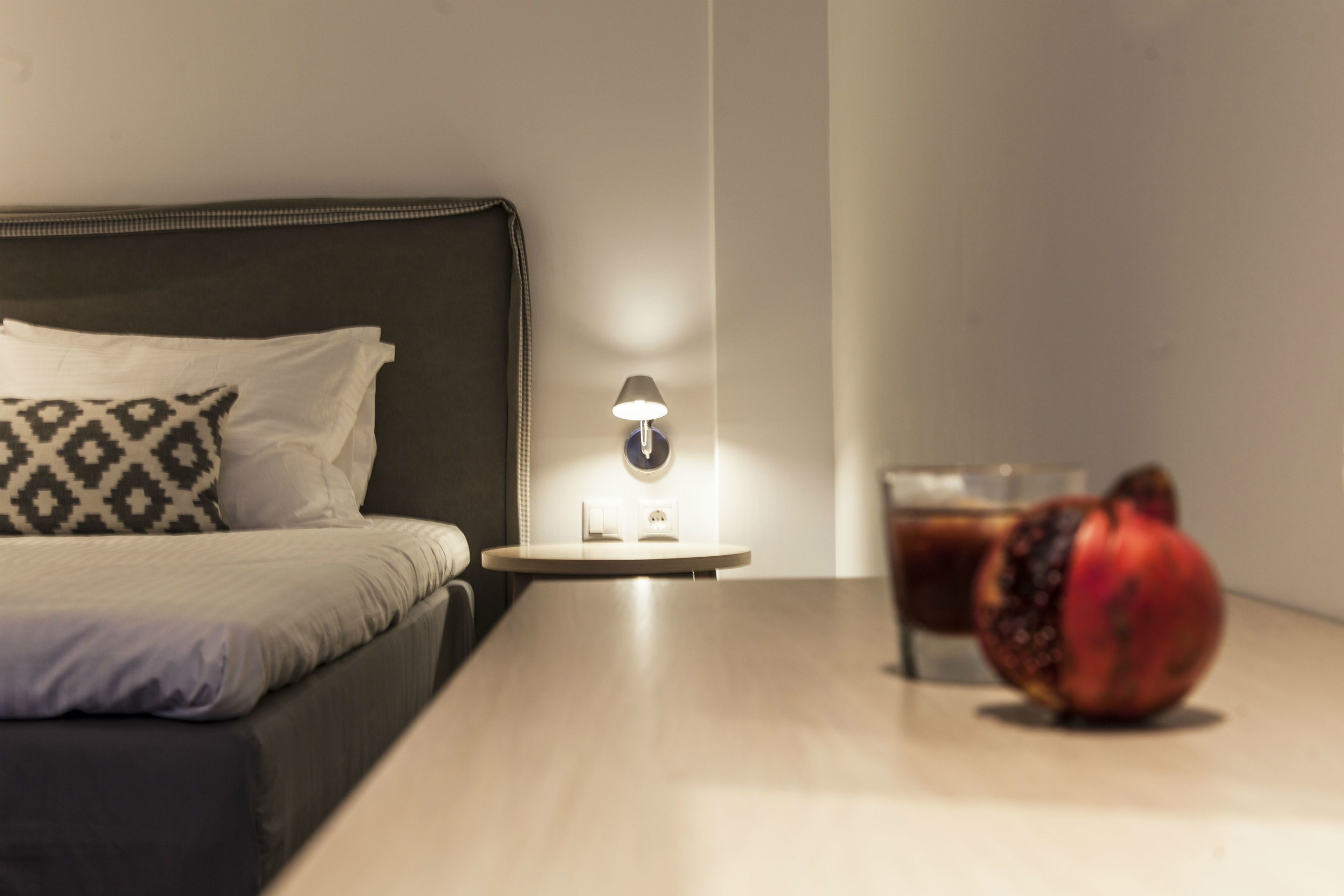 A neatly made bed with patterned pillows, a bedside lamp, a glass of a dark beverage, and a pomegranate on a wooden table.