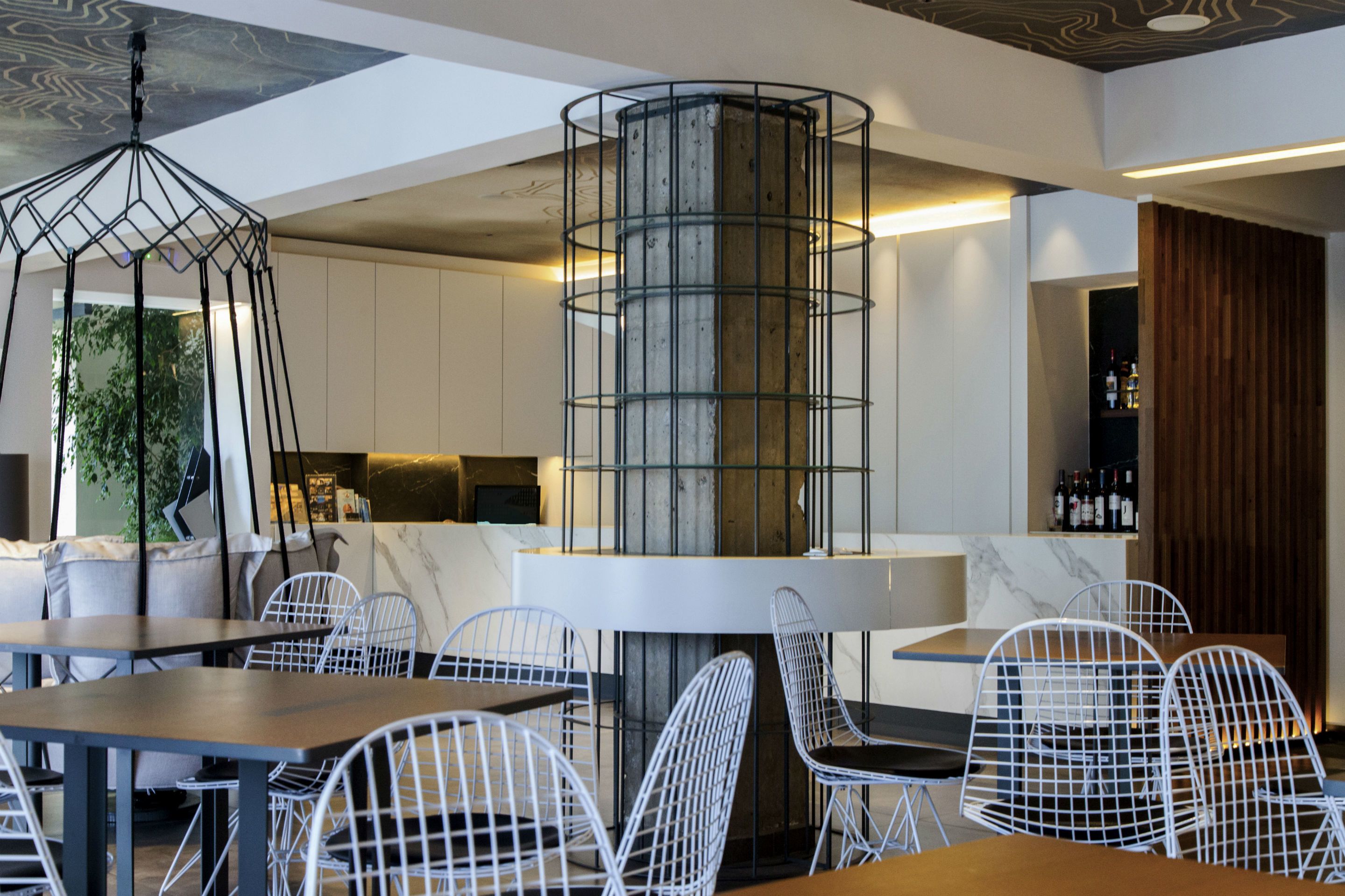 Un interior de restaurante moderno con sillas de metal blanco, una columna central de madera y una zona de bar con botellas.