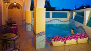 Zona de piscina con una persona en el agua, rodeada de macetas con flores y un espacio de descanso con sillas y mesas.