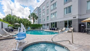 Una piscina con sillones reclinables y un jacuzzi, rodeada por un edificio con amplias ventanas y una terraza pavimentada.