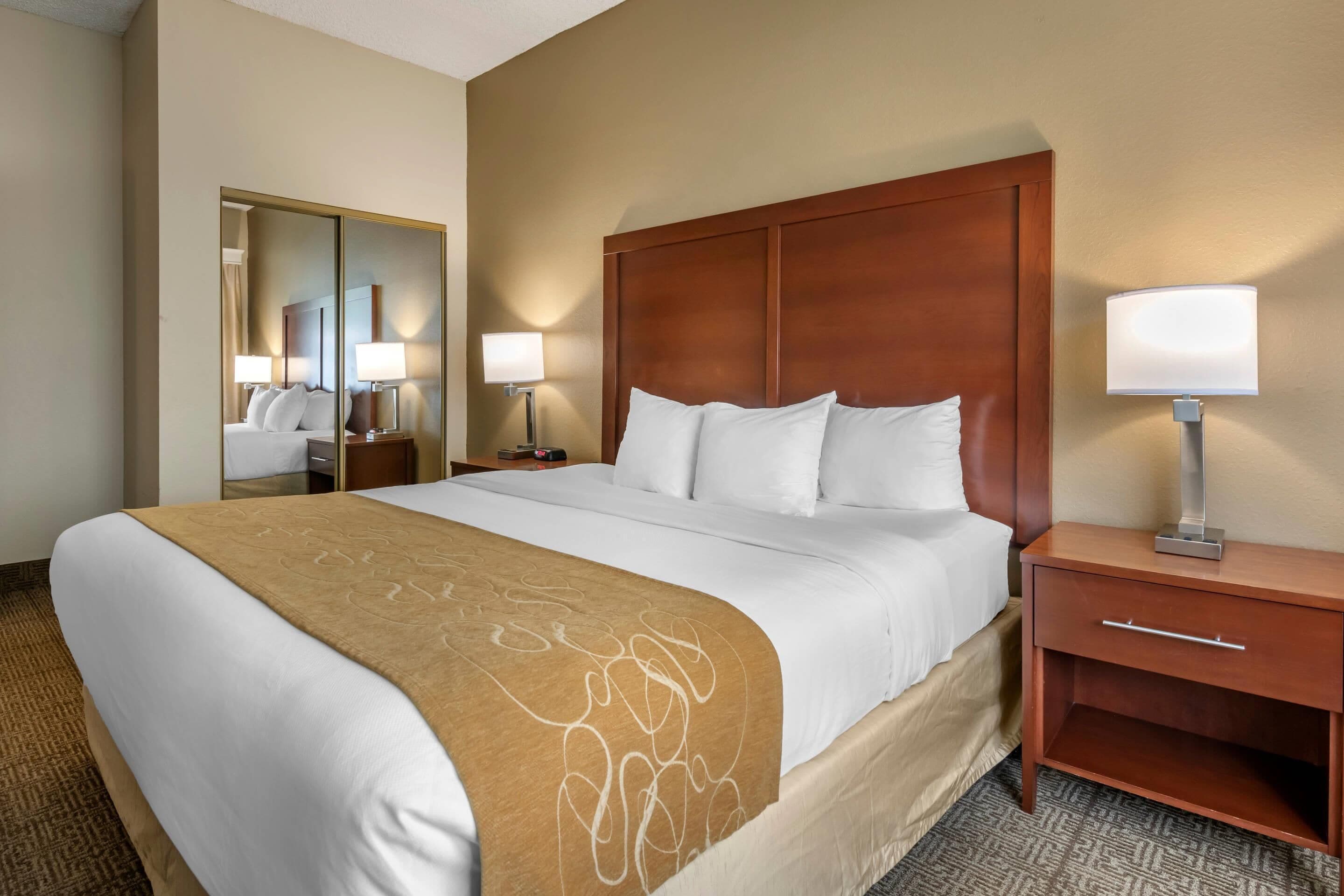 Habitación de hotel con una cama grande, cabecera de madera, mesita de noche con lámpara y espejo.