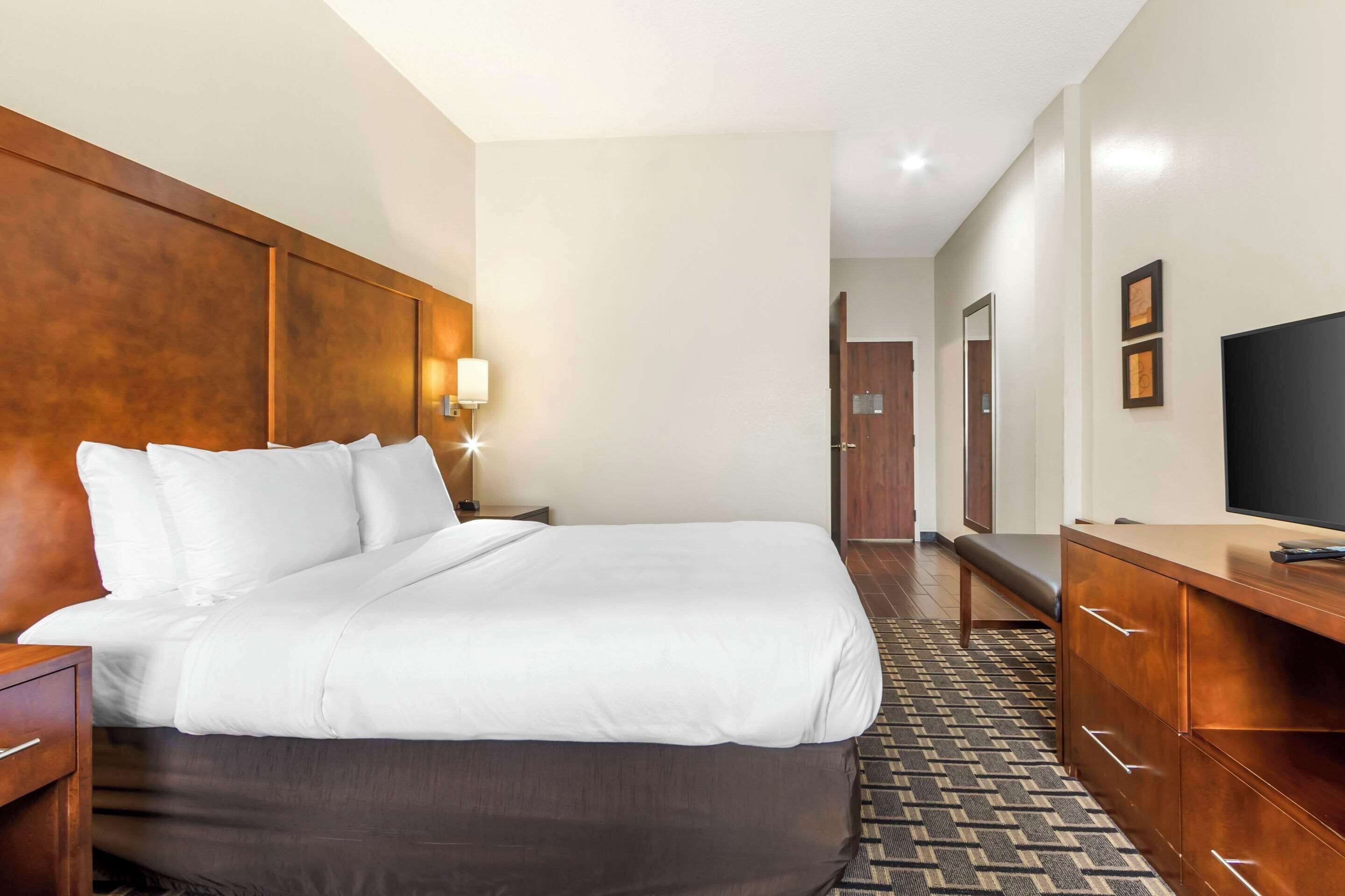 A hotel room with a large bed, wooden headboard, bedside table, lamp, and a TV on a wooden dresser.