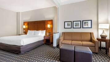 A hotel room with a large bed, a sofa, an ottoman, a nightstand, a lamp, and framed pictures on the wall.