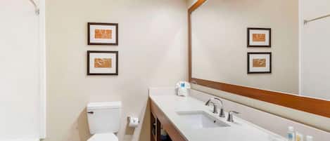 Un baño moderno con lavamanos, espejo y retrete blancos. Se observan cuadros enmarcados en la pared y un perchero para toallas.