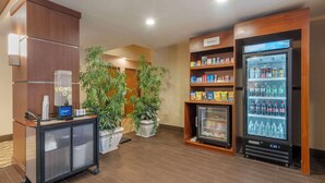 A hotel lobby with a self-service beverage dispenser, a cart with a coffee maker, and potted plants.