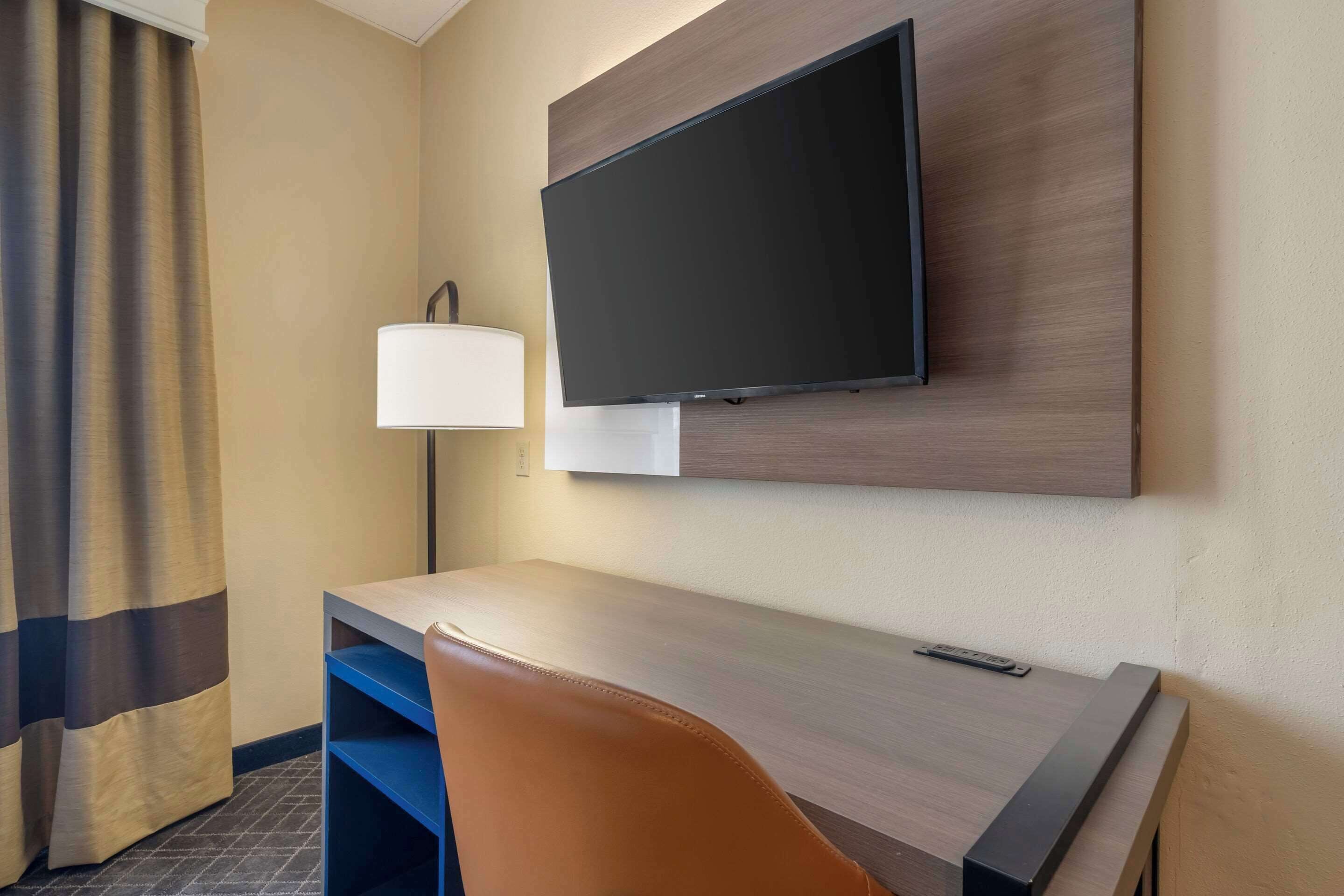 A hotel room with a flat-screen TV mounted on the wall, a desk with a chair, a lamp, and a remote control on the desk.