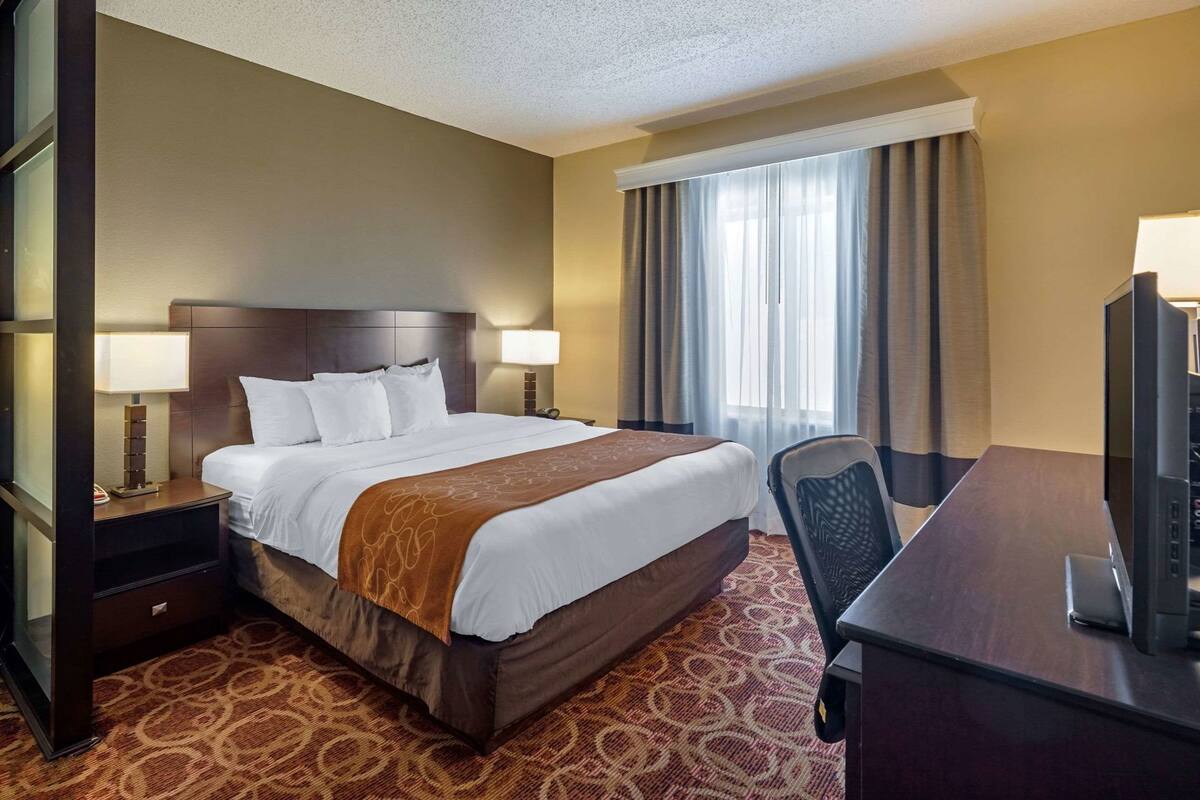 A hotel room with a large bed, a desk with a television, a nightstand, and a window with curtains.