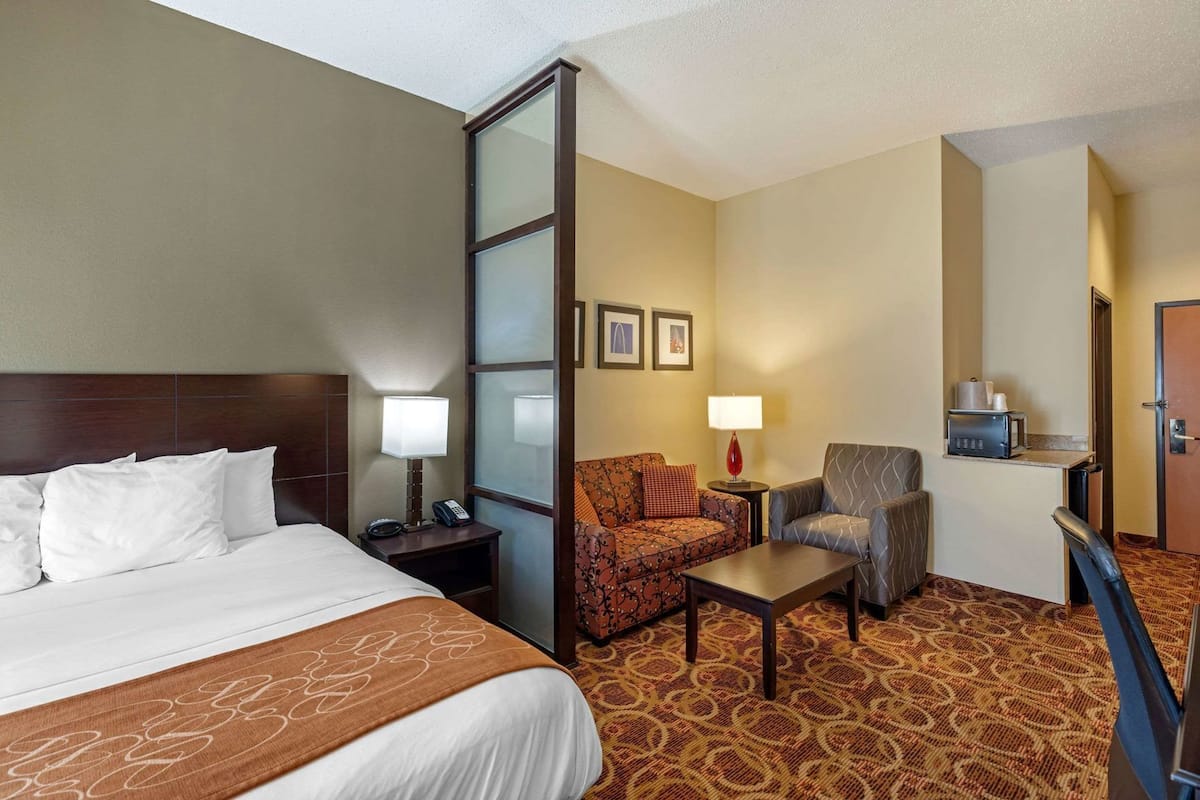A hotel room with a bed, a desk, a chair, a small table, a lamp, a microwave, and a door.