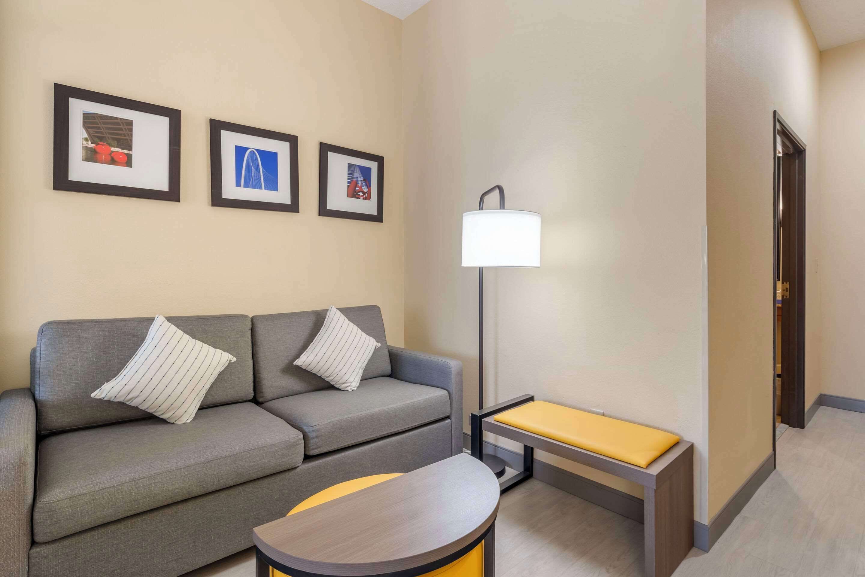 Quarto de hotel moderno com sofá cinzento, uma mesa pequena e um banco. Há três quadros na parede.