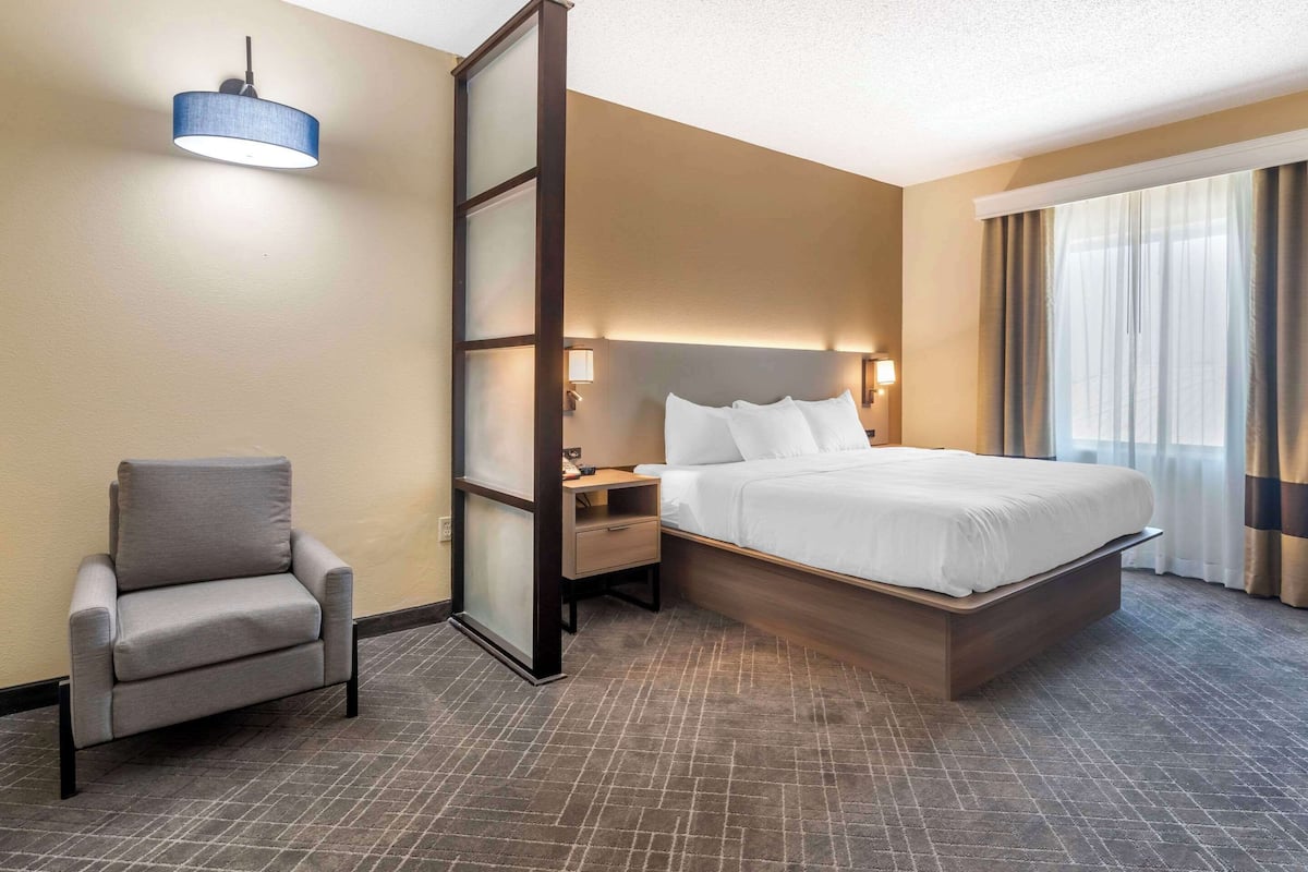 A hotel room with a bed, a chair, a nightstand, a lamp, and a large window with curtains.
