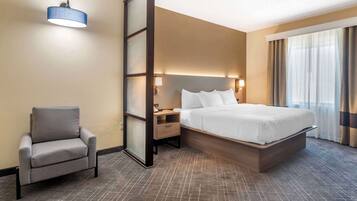 Ein Hotelzimmer mit Bett, Sessel, Nachttisch, Lampe und einem großen Fenster mit Vorhängen.