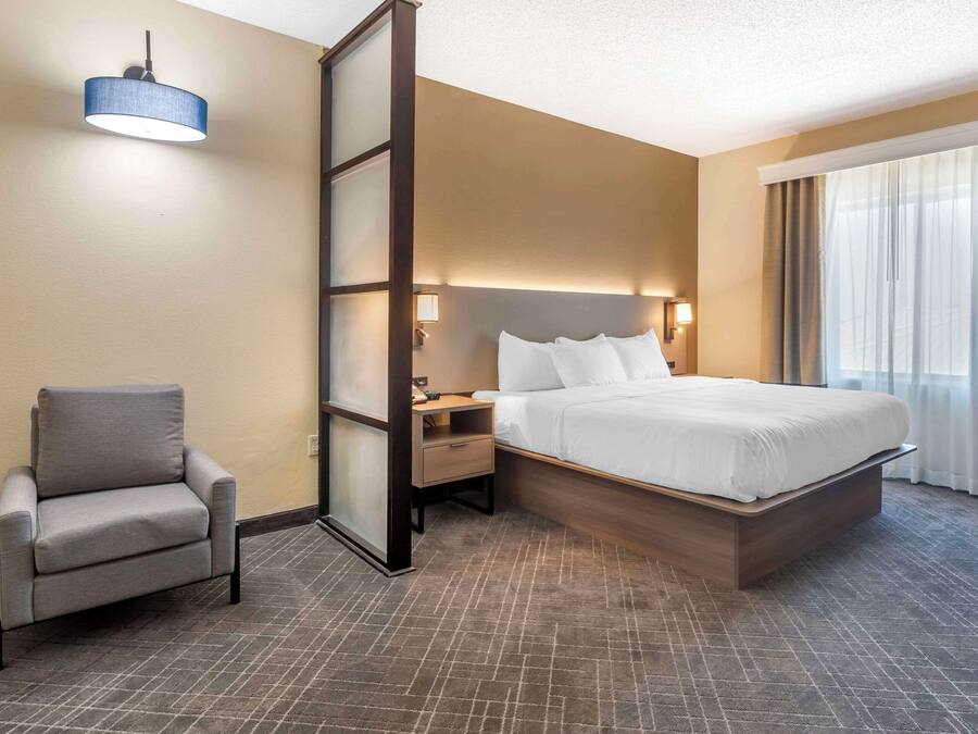 Ein Hotelzimmer mit Bett, Sessel, Nachttisch, Lampe und einem großen Fenster mit Vorhängen.
