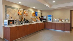 A modern hotel kitchen with a self-service breakfast bar, microwave, coffee machines, and a variety of food items.