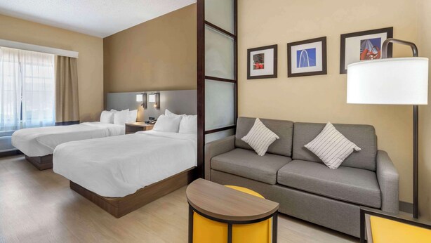A hotel room with two beds, a sofa, a small table, and a lamp. There are framed pictures on the wall.