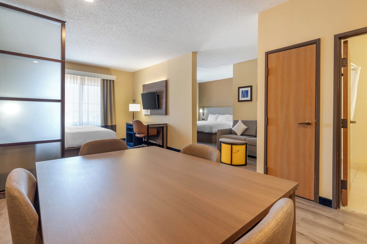 A hotel room with a wooden dining table, a sofa, a bed, a TV, and a desk with a lamp.