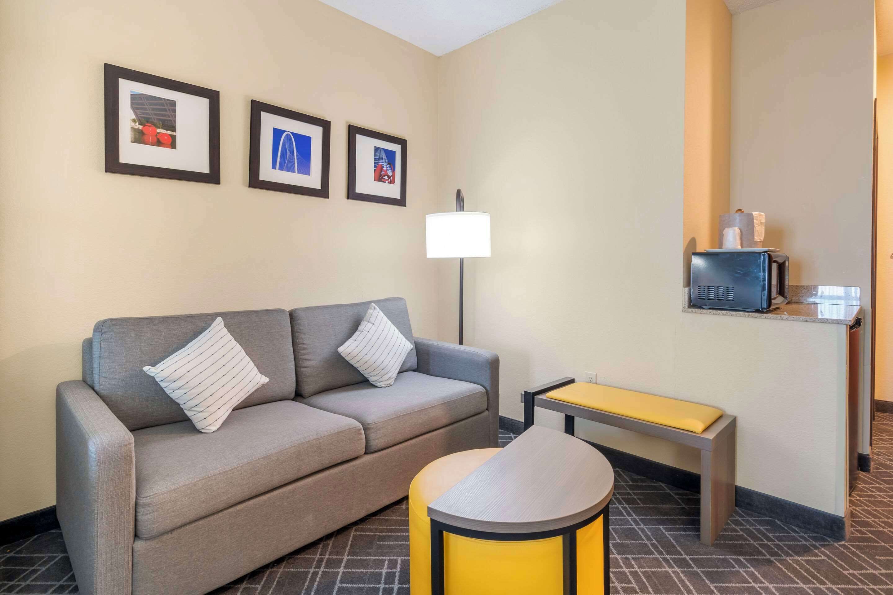 A hotel room with a grey sofa, a small yellow table, a lamp, and framed pictures on the wall.