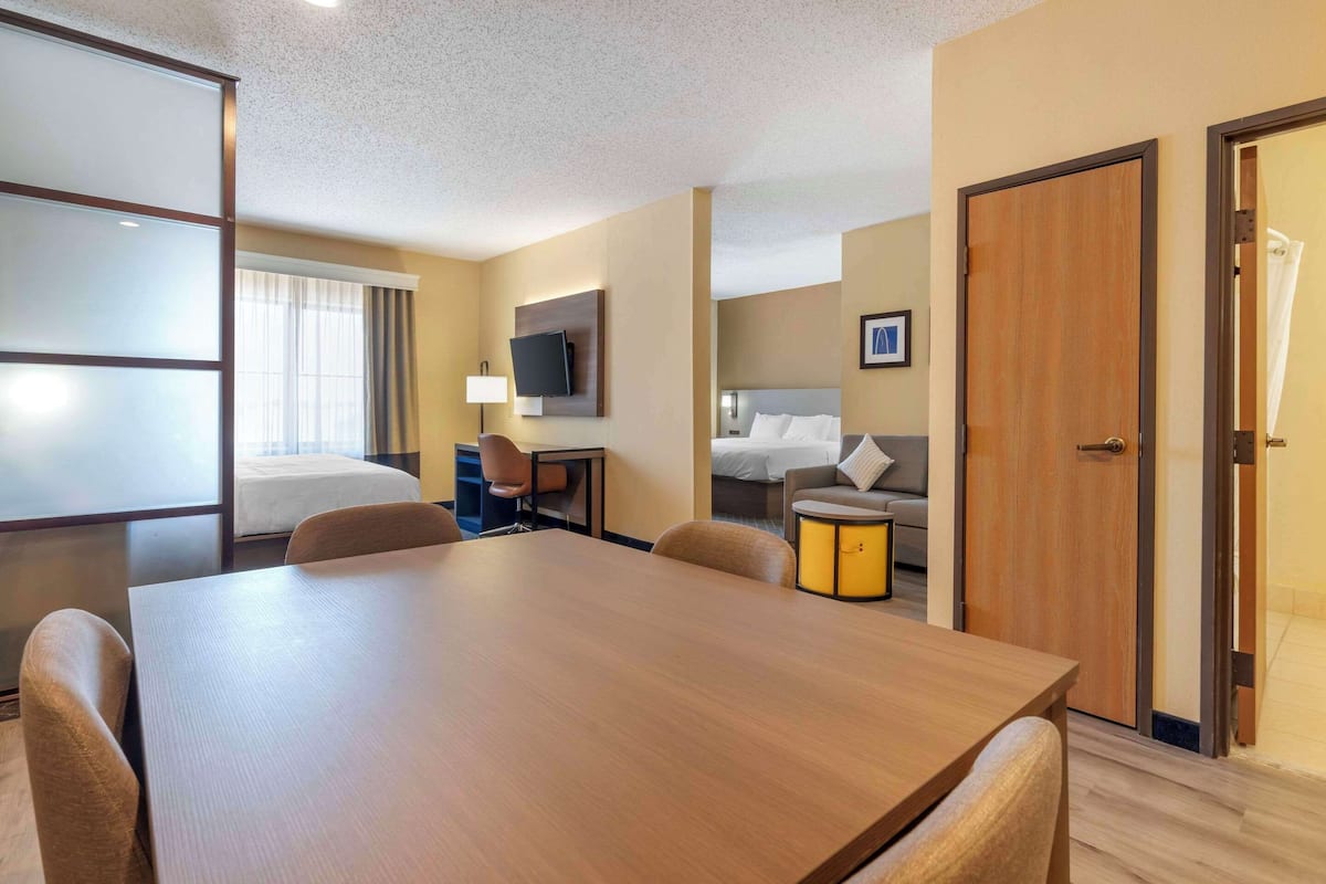 A hotel room with a wooden dining table, a sofa, a bed, a TV, and a door.