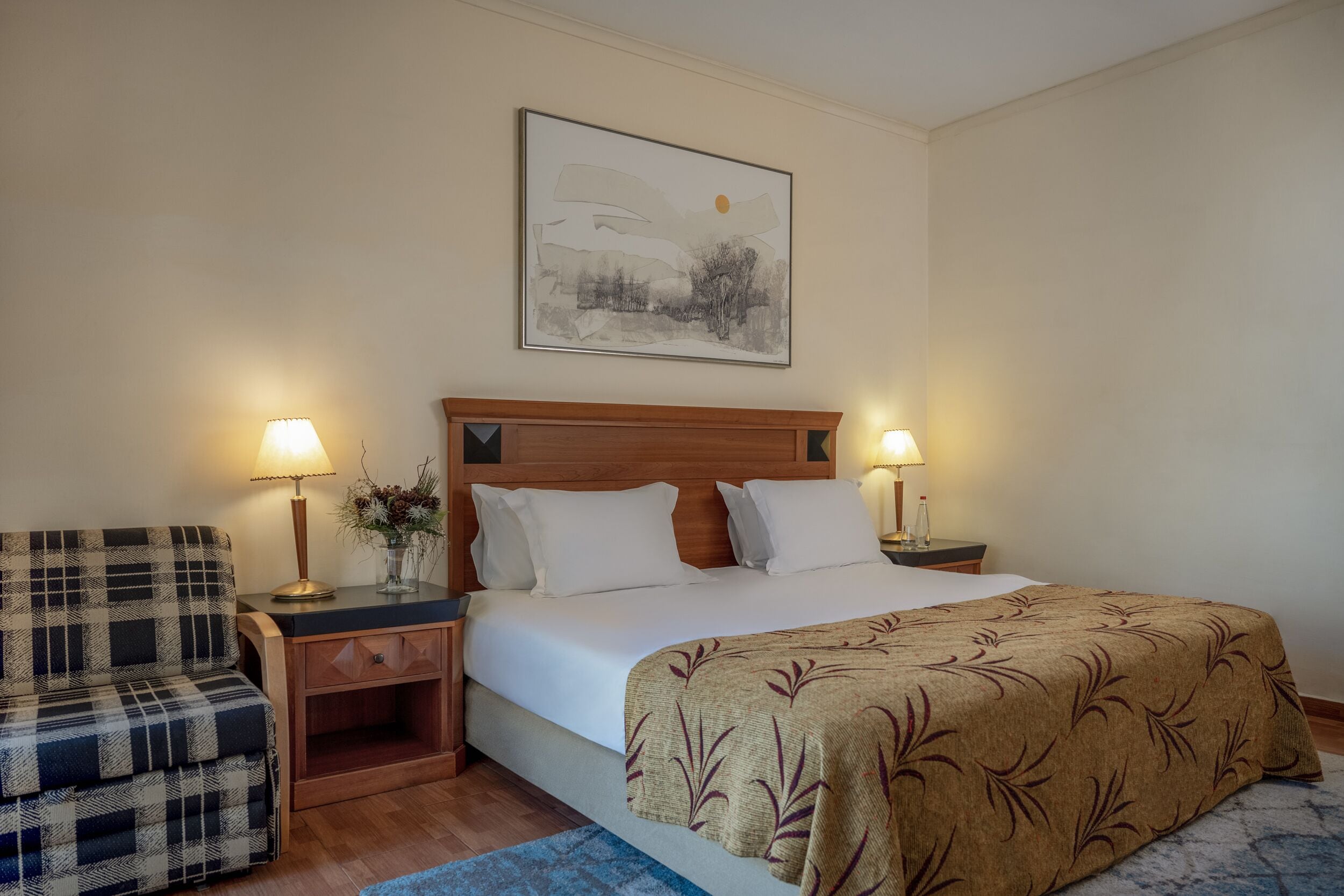 A hotel room with a bed, two bedside tables with lamps, a wooden headboard, a plaid armchair, and a framed picture on the wall.