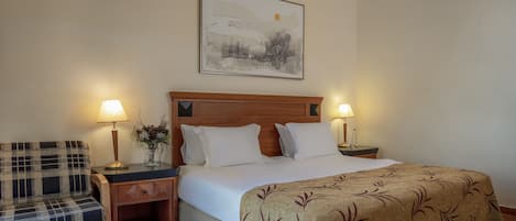 A hotel room with a bed, two bedside tables with lamps, a wooden headboard, a plaid armchair, and a framed picture on the wall.