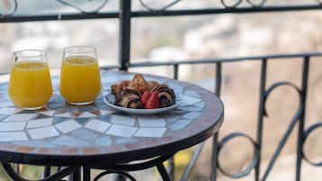Una mesa con dos vasos de jugo de naranja y un plato de comida, que incluye fresas y cruasanes, en un balcón con barandilla de metal y vistas a montañas lejanas.